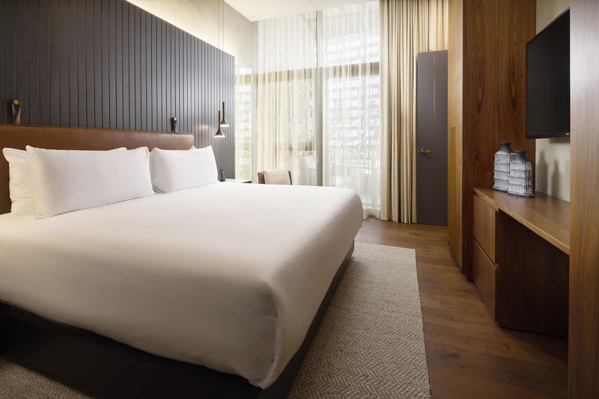 A hotel room with a large bed, wooden headboard, a flat-screen TV, a wooden wardrobe, and a large window with sheer curtains.