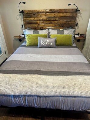 Uma cama com colcha listrada em cinza e branco, um travesseiro com a palavra 'Relax' e cabeceira de madeira.