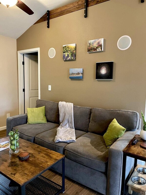A living room with a gray sofa, green and white throw pillows, a coffee table with a glass container of apples, and a small wooden table with a bowl and a remote control.