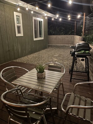 An outdoor patio with a wooden table and chairs, a grill, and string lights.