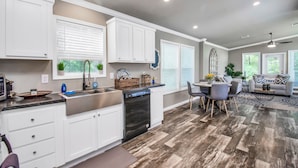 A modern kitchen with white cabinets, a dishwasher, and a dining area with a gray sofa and a coffee table. - The Lantana Tiny Home  ~ Cottage in the Woods ~ NEW HOT TUB & DOGGY PLAY YARD (Willis)