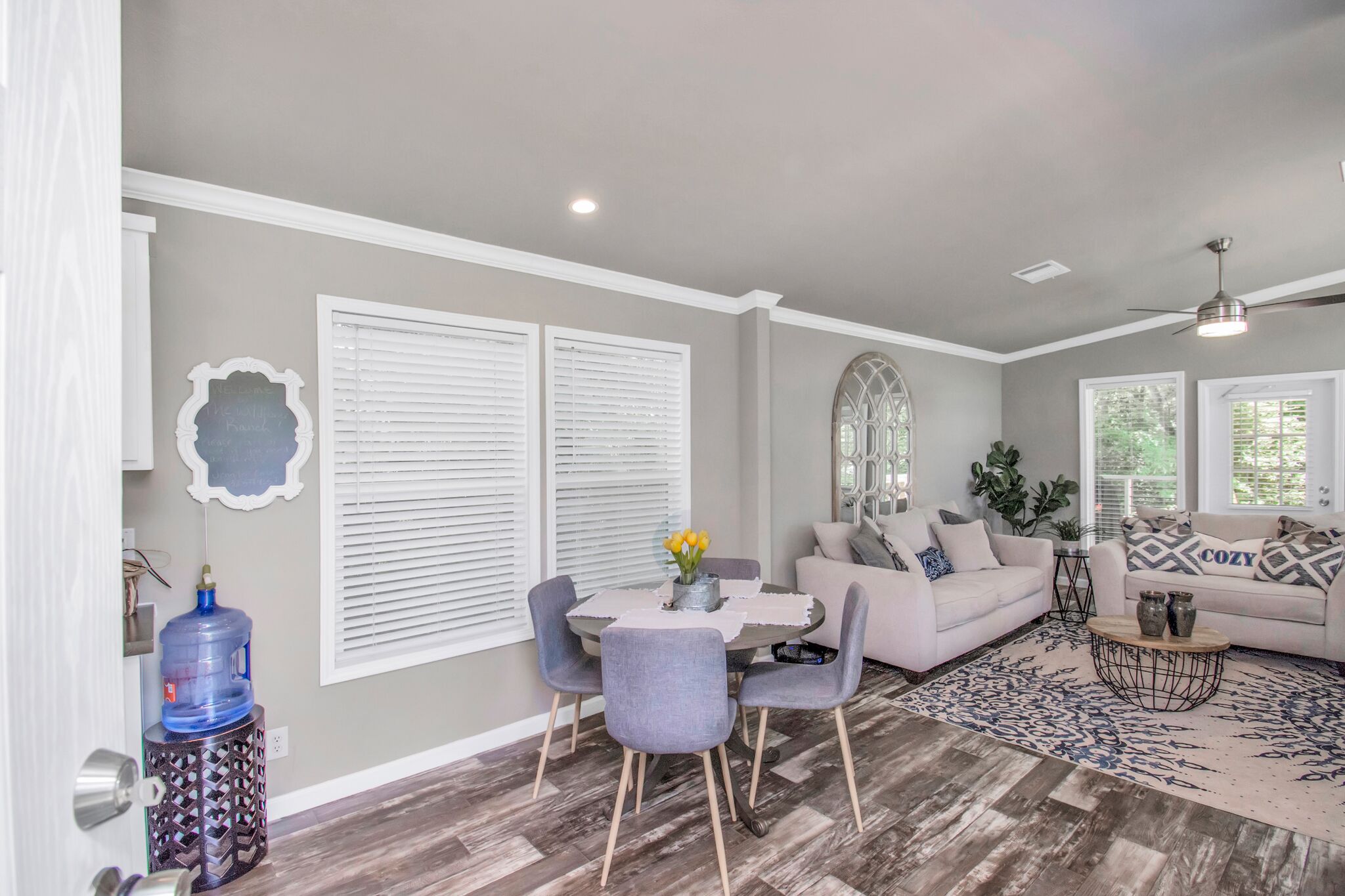 A modern living room with a dining area, featuring a large water dispenser, grey upholstered chairs, a white sofa, and a patterned rug.