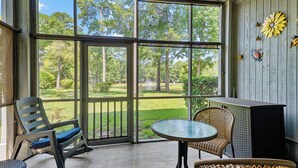 A covered patio with a view of a lawn and trees, furnished with a rocking chair, a table, and chairs.