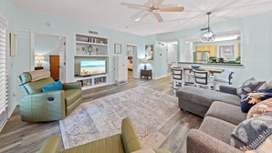 A spacious living room with a ceiling fan, a large rug, and a view of the beach through the open door.