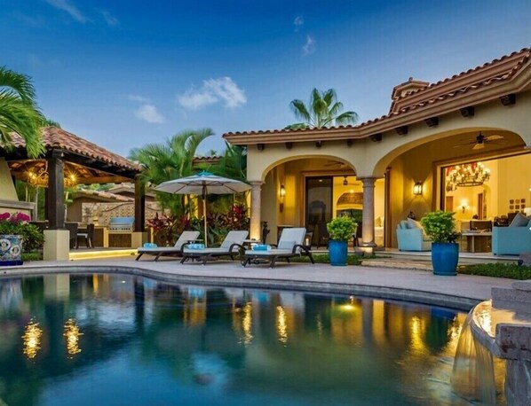 A pool area with lounge chairs and umbrellas, a covered patio, and a well-lit interior.