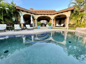 A pool area with lounge chairs and umbrellas, a covered patio with seating, and a well-maintained garden with palm trees.