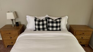 Una cama bien hecha con sábanas blancas, dos mesitas de noche de madera con cajones, una lámpara y una cafetera sobre una de ellas.