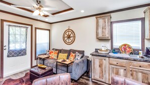 A modern living room with a gray sofa, a wooden dining table, and a kitchen area with wooden cabinets and a marble countertop. - Bluebonnet Tiny Home ~ HOT TUB (Willis)