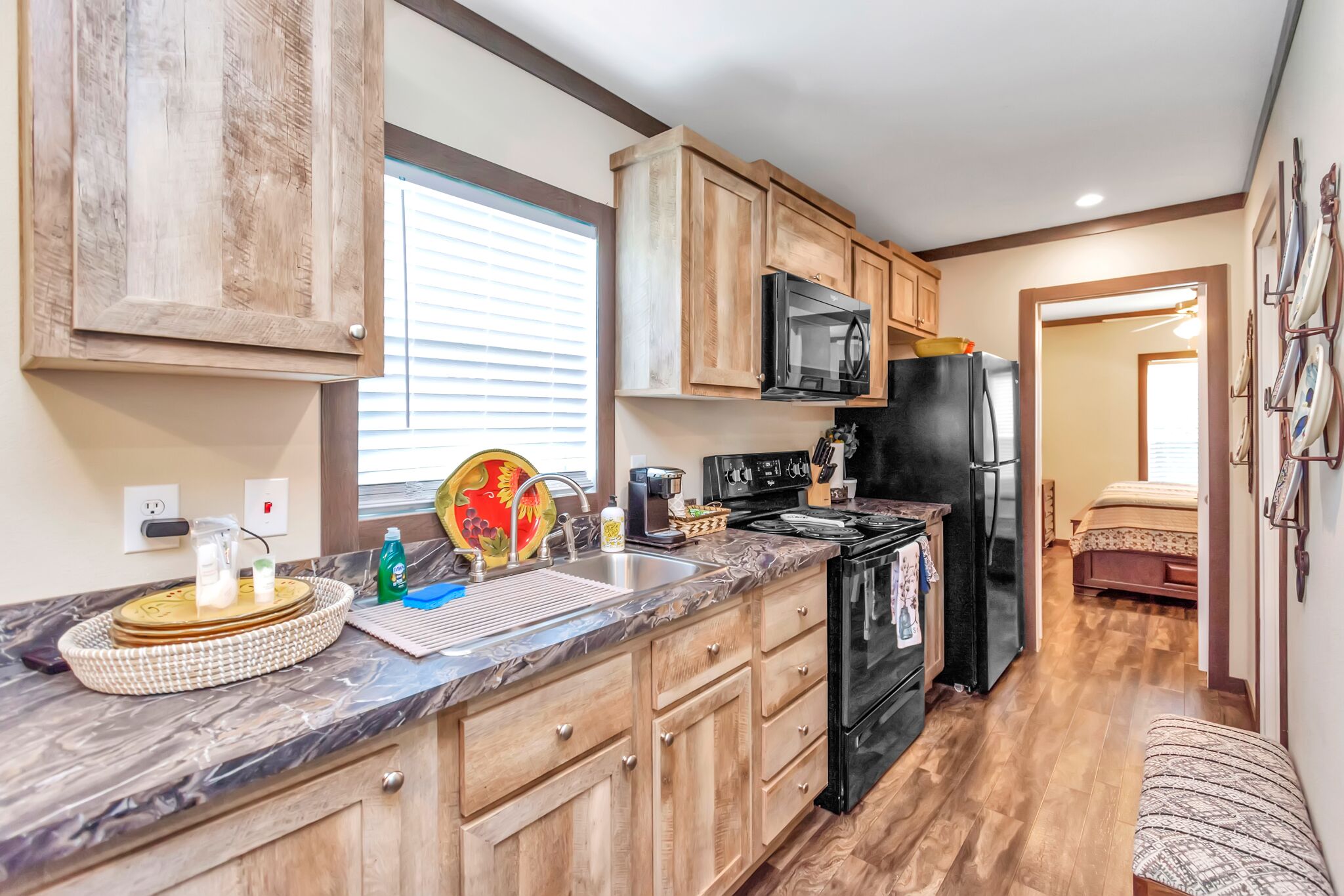 A compact kitchen with wooden cabinets, a black refrigerator, a microwave, and a stove. There is a sink with a dish rack and a window above it.