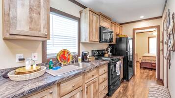 A compact kitchen with wooden cabinets, a black refrigerator, a microwave, and a stove. There is a sink with a dish rack and a window above it.