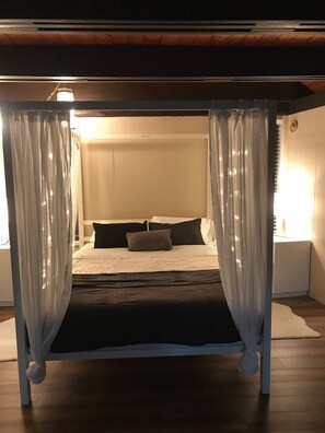 Una cama con dosel con cortinas traslúcidas, un cabecero blanco y una mesita de noche con una lámpara.