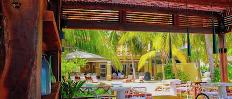 A covered outdoor seating area with tables and chairs, a pool, and palm trees in the background.