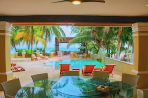 Een tafel met glazen blad, rieten stoelen, een zwembad en ligbedden met uitzicht op de oceaan en palmbomen.