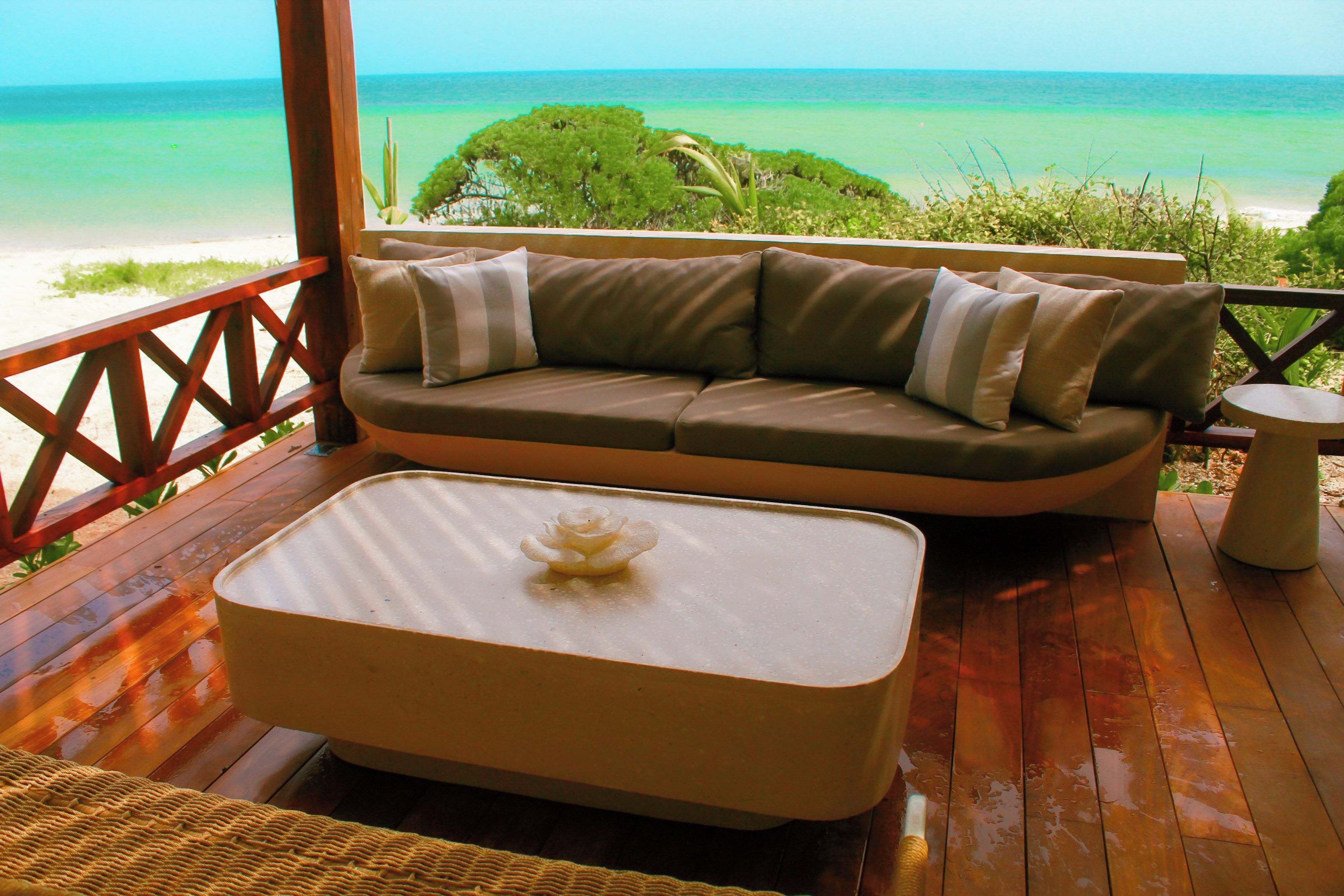 A wooden deck with a beige sofa and a white table, overlooking a beach and ocean.