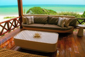 A wooden deck with a beige sofa and a white table, overlooking a beach and ocean. - Esciros Luxury Villa (Progreso)