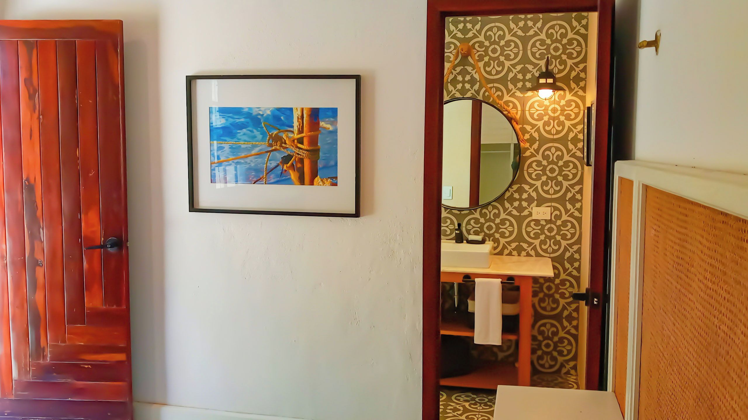 A framed painting of a sailboat on a wall, a wooden door, and a bathroom with a mirror and patterned wallpaper.