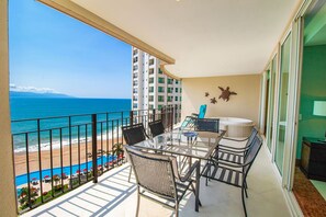 A balcony with a glass table and chairs, a hot tub, and a view of the beach and ocean.