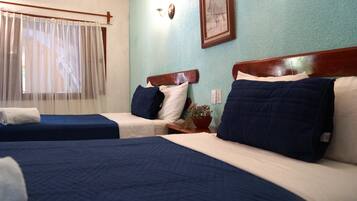 Habitación de hotel con dos camas, cada una con ropa de cama azul y almohadas blancas. Se ve una ventana con cortinas translúcidas, una lámpara de pared y un cuadro enmarcado sobre las camas.