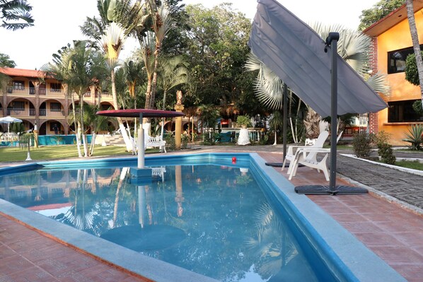 Una piscina con sillones de descanso, un área de estar cubierta y un edificio de techo rojo al fondo.