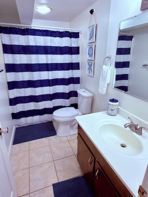 A bathroom with a striped shower curtain, a toilet, a sink with a mirror, and a towel rack. - Blue & White Getaway - 5 min to Beaver Stadium (State College)