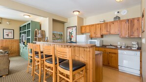 A kitchen with wooden cabinets, a white refrigerator, a microwave, and a coffee maker. There are three wooden bar stools with black cushions. - Large Corner Condo in Creekside with a Patio to enjoy the Views  (Silver Star Mountain)
