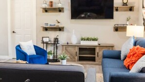 Een woonkamer met een aan de muur bevestigde televisie, een blauwe fauteuil, een grijze bank en een houten tv-meubel met planken.