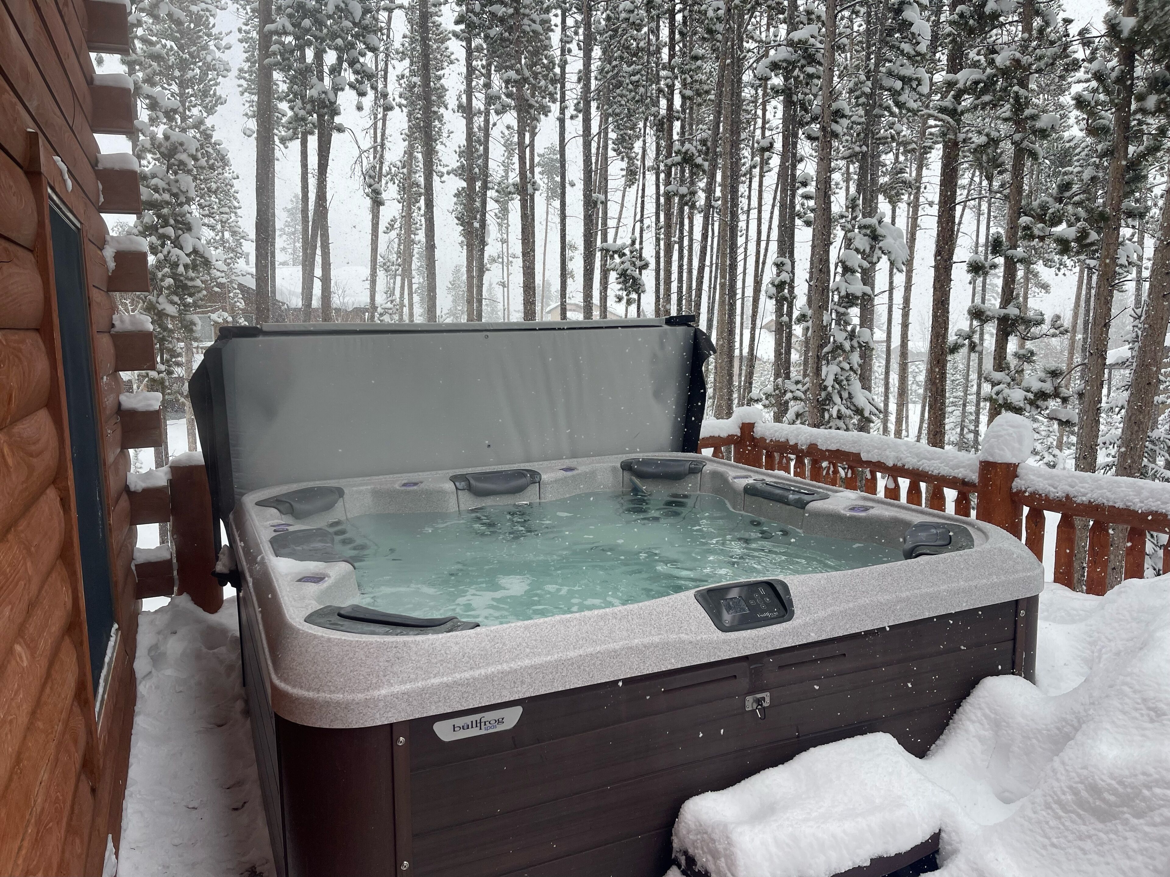 A hot tub with a wooden deck and a snowy forest background.