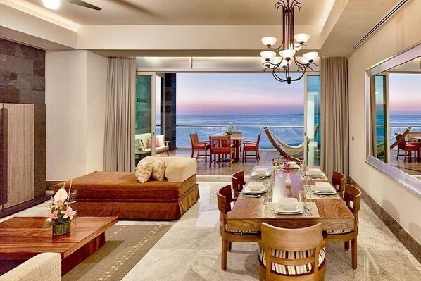 A modern hotel room with a dining area, a sofa, a view of the ocean, and a large window.