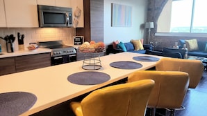 A modern kitchen with a central island, a microwave, and a stove. There is a dining area with yellow chairs and a table. The room has a brick wall and a window with a view. - Rustic Realm/2BR2Bath/KayBaileyCenter/Pool/GYM (Dallas)