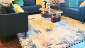A living room with two sofas, a coffee table, and a rug. There is a painting on the wall and a lamp next to a window. - Rustic Realm/2BR2Bath/KayBaileyCenter/Pool/GYM (Dallas)