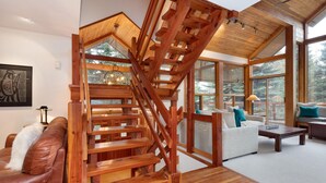 A wooden staircase leading to a loft area with a large window, a sofa, and a coffee table in a modern living room. - Luxury 5 Bedroom Blueberry Chalet (Whistler)