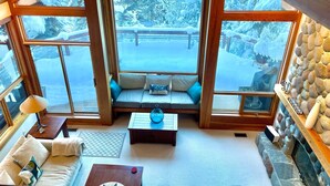 A cozy room with a large window overlooking a snowy landscape, a stone fireplace, and a comfortable seating area. - Luxury 5 Bedroom Blueberry Chalet (Whistler)