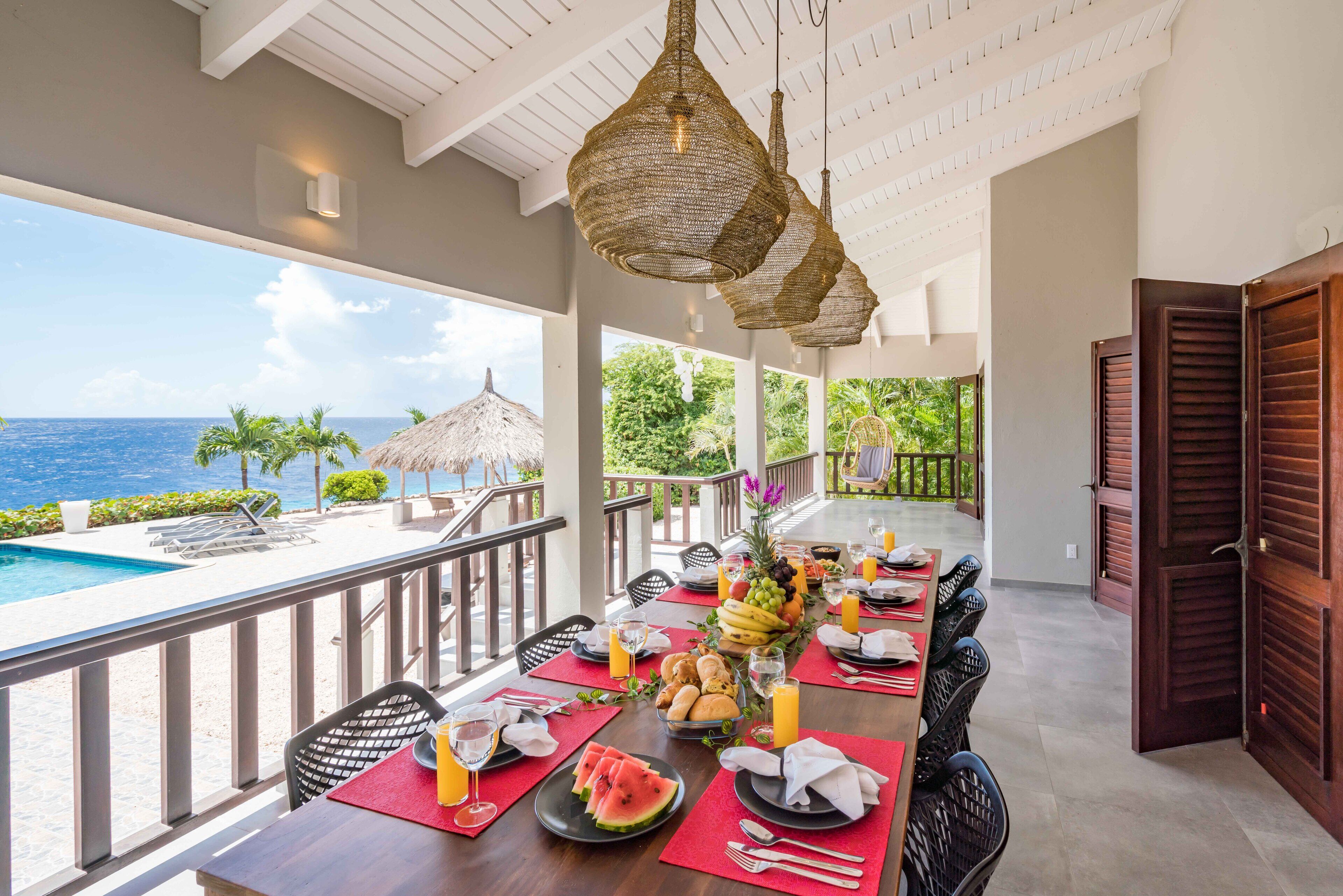 A dining area with a view of the ocean, a pool, and a thatched-roof structure.