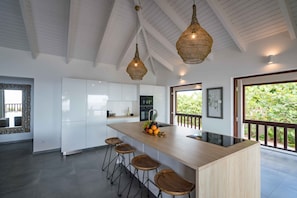 A modern kitchen with a central island, bar stools, hanging wicker pendant lights, and a view of greenery through a large window. - Villa Seven - Coral Estate, Spectacular Views - Ocean Access ! (Curaçao)