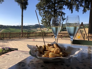 Una mesa con una porción de papas fritas y dos copas de vino blanco, ubicada en una terraza con vista a una valla de madera y árboles.