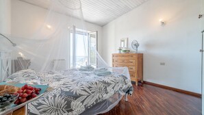 Quarto com cama coberta por um edredom estampado com flores, cômoda de madeira, mesa de cabeceira com abajur e janela com vista para o exterior.