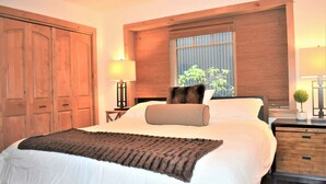 Un dormitorio con una cama grande, armario de madera, mesitas de noche con lámparas y una ventana con vistas a la vegetación.