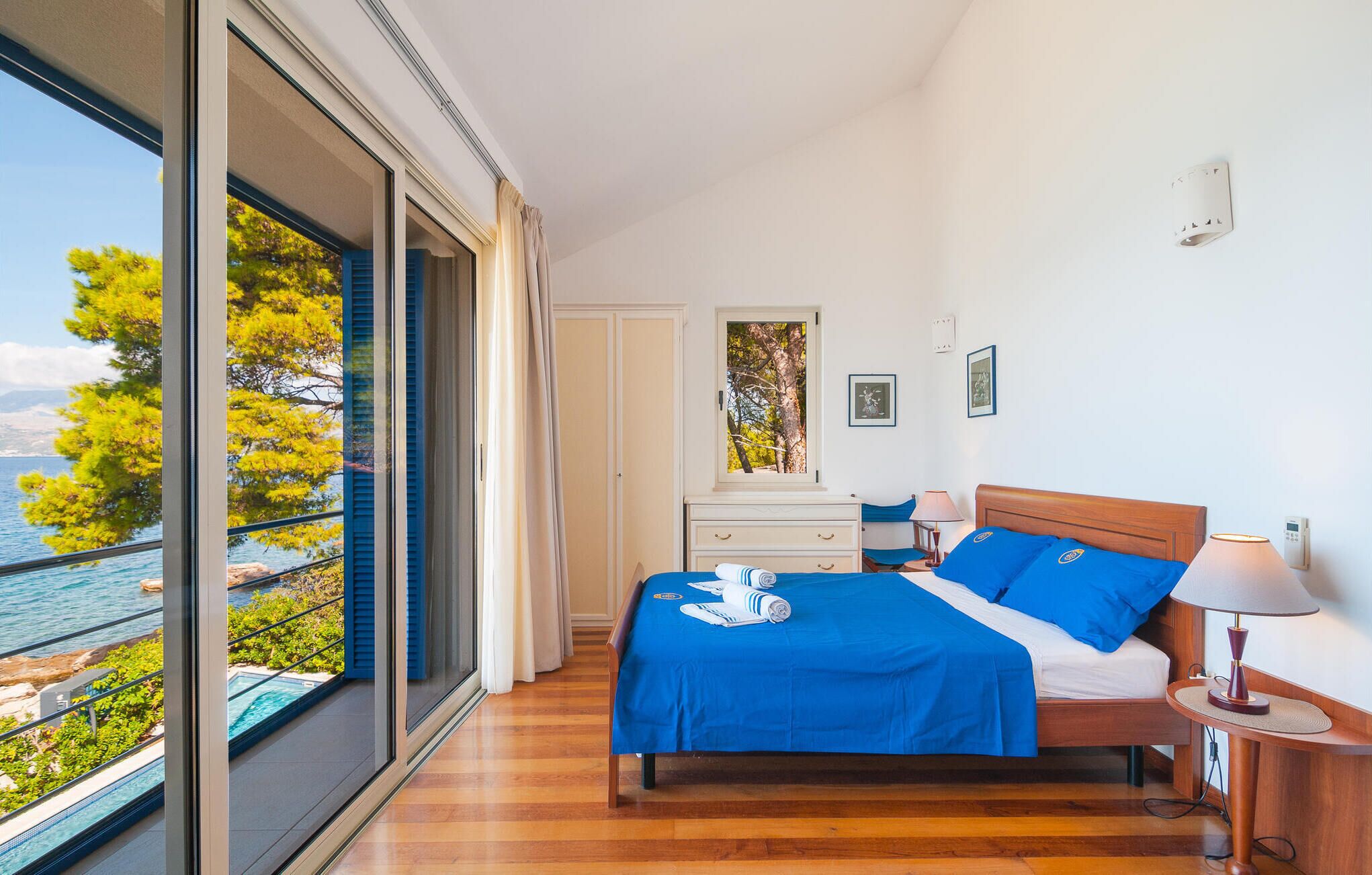 Ein Schlafzimmer mit großem Fenster, einem Bett mit blauer Bettwäsche, einem Nachttisch mit Lampe und Blick aufs Meer.
