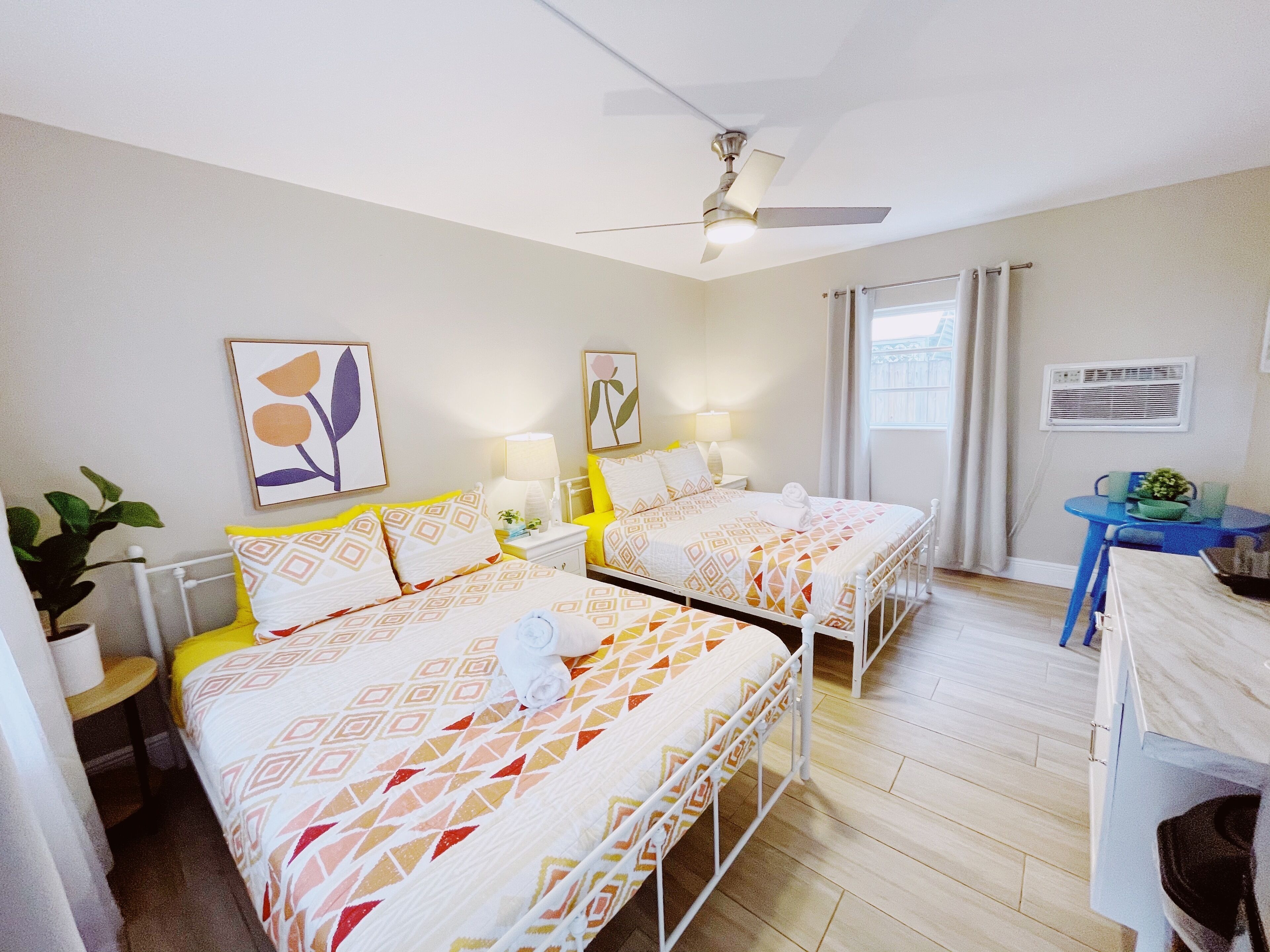 Een hotelkamer met twee bedden, een plafondventilator, een raam met gordijnen, een kleine tafel met een plant en een airconditioning aan de muur.