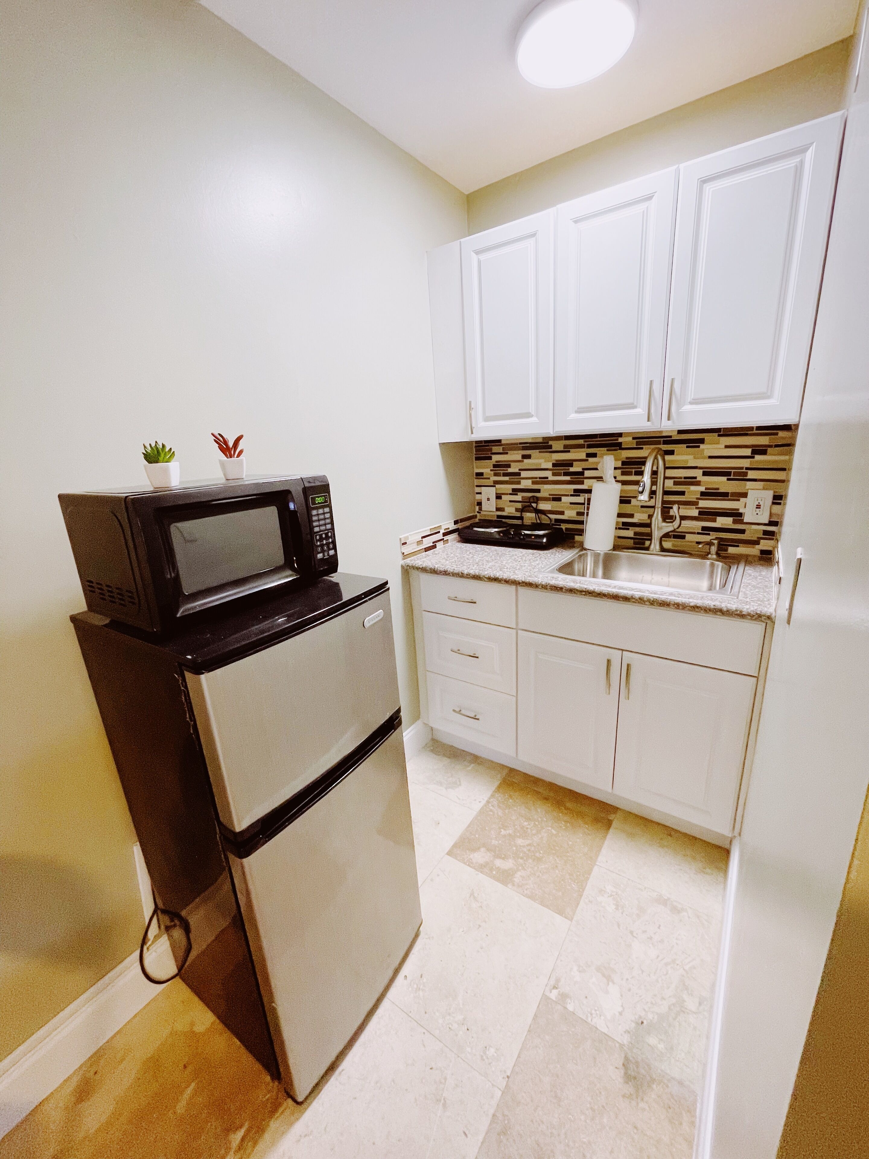 A compact kitchen with white cabinets, a stainless steel refrigerator, a microwave, and a tiled backsplash.