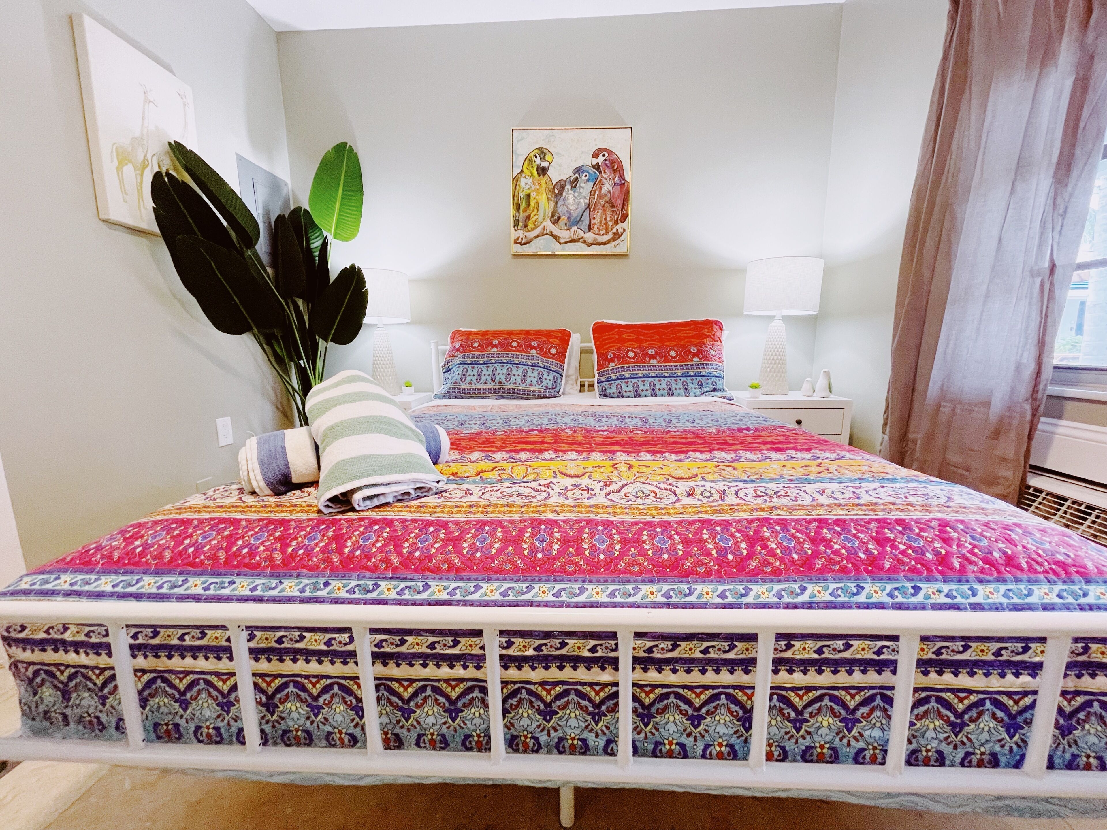 A bedroom with a colorful bedspread, a large plant, and a painting on the wall.