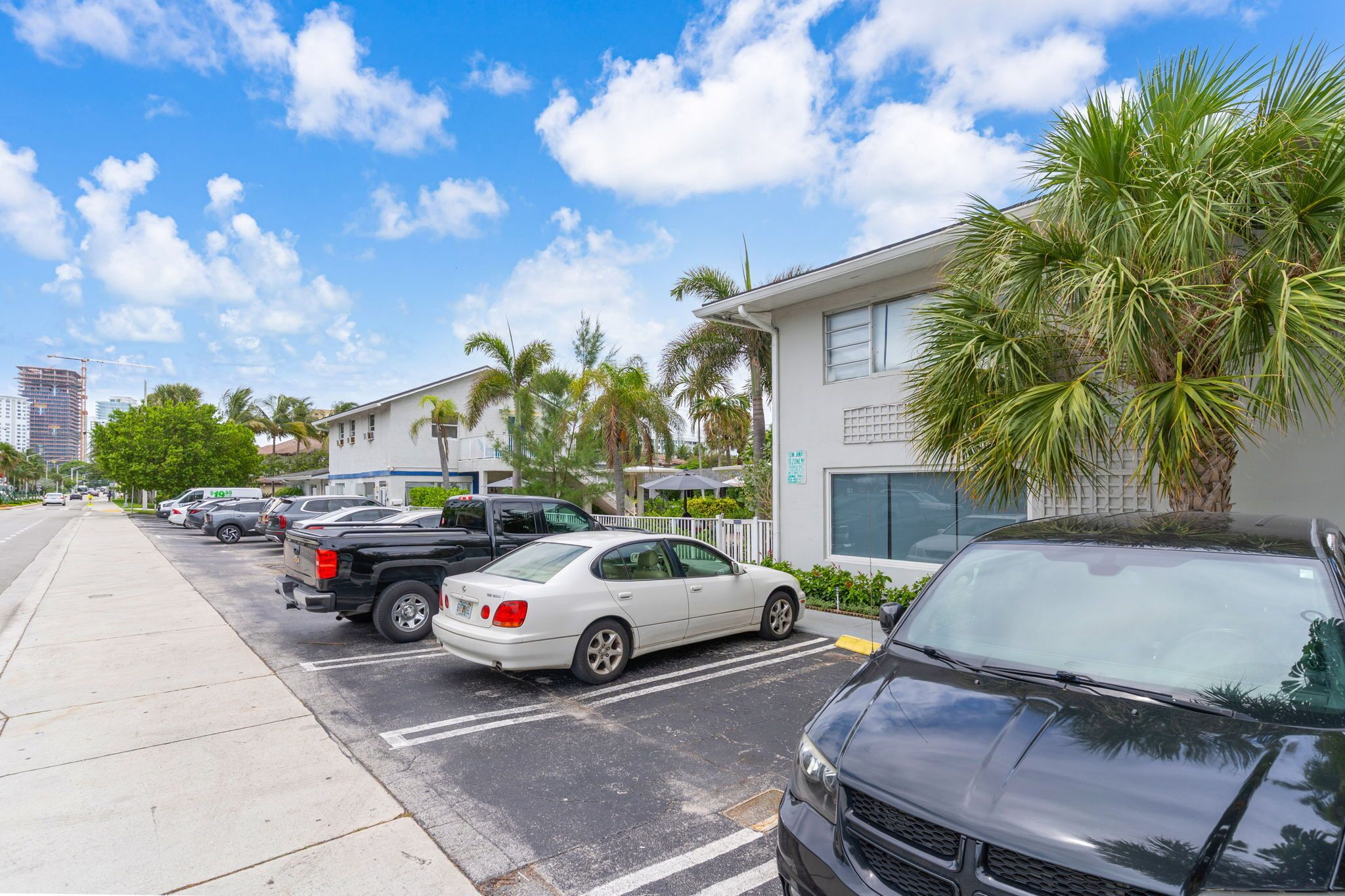 A parking lot with several cars, a building with a white facade, and palm trees.