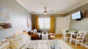 Una habitación luminosa con una cama, un sofá, una mesa de comedor con sillas, un ventilador de techo y un televisor en la pared.