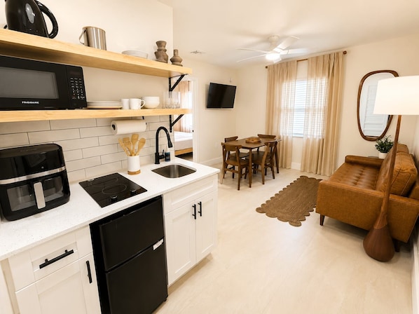 A compact kitchen with a microwave, coffee maker, and a refrigerator. There is a dining area with a table and chairs, and a sofa in the living room. - Sea Steps by Lowkl (Pompano Beach)