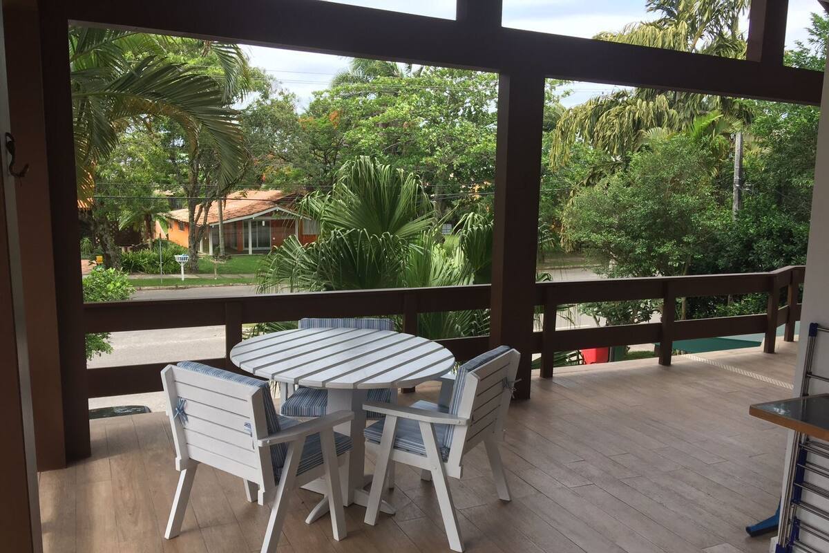 A covered outdoor area with a table and chairs, a wooden deck, and a view of a residential area with trees and a house.