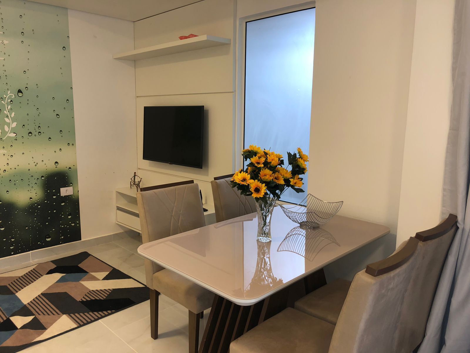 A modern living room with a flat-screen TV, a dining table with chairs, a vase of sunflowers, and a geometric rug.