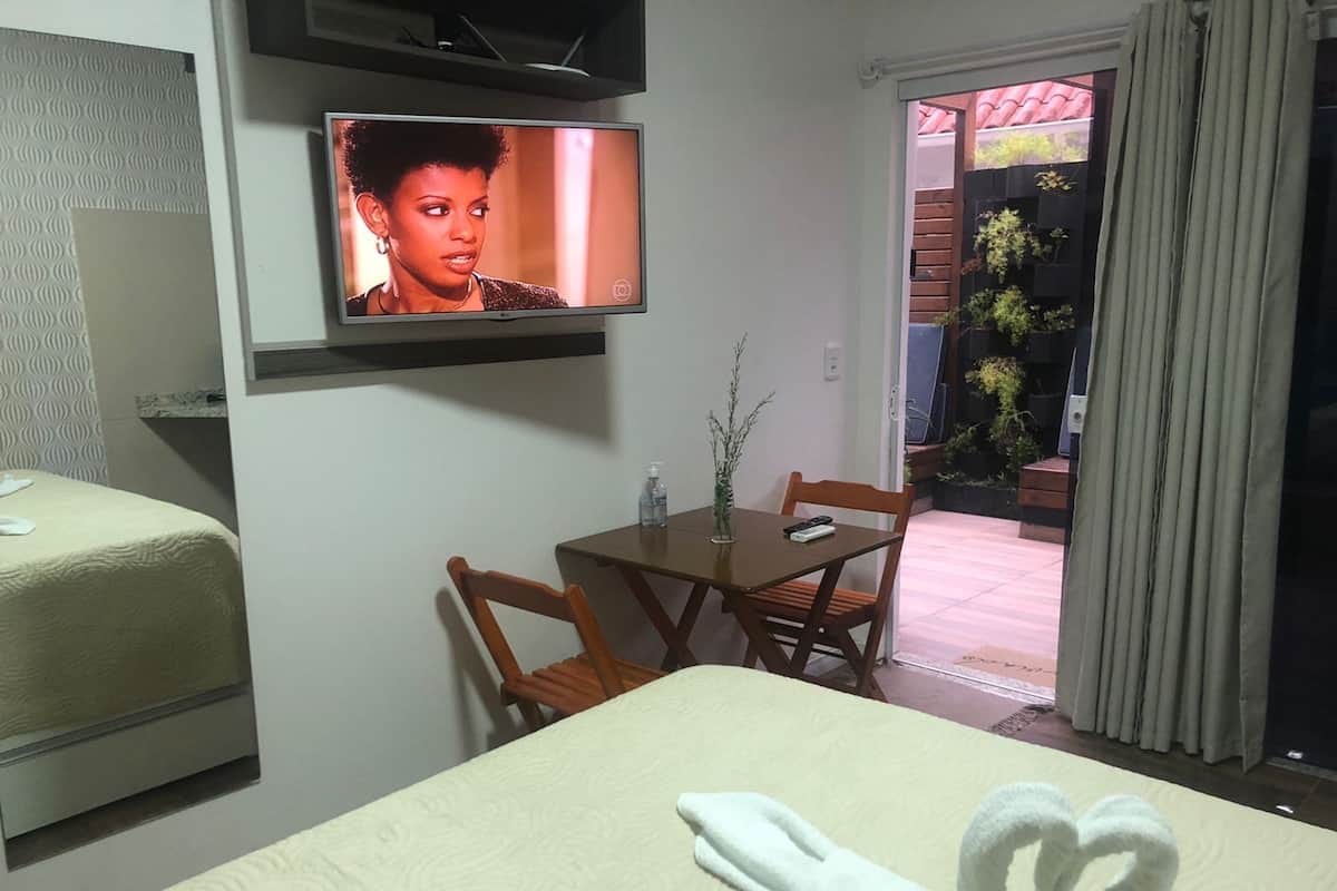 A hotel room with a bed, a TV mounted on the wall, a small table with chairs, and a view of the outside through a door.