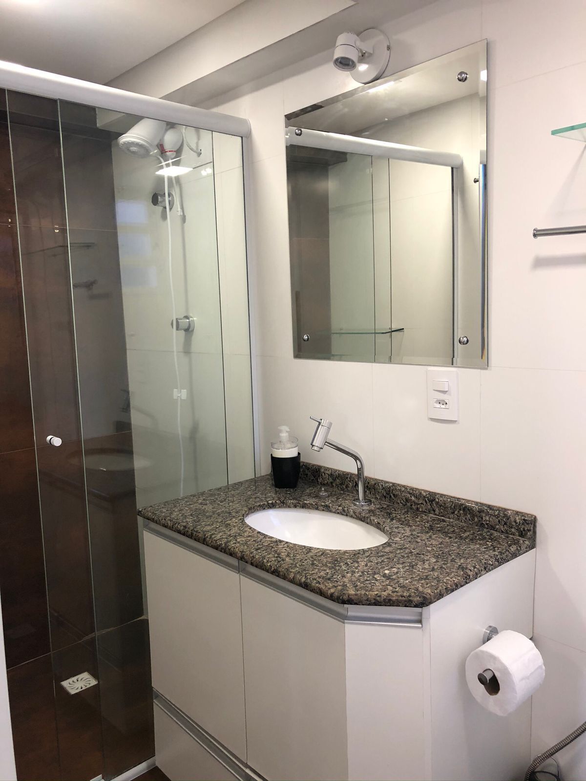 Un baño moderno con cabina de ducha de vidrio, lavamanos con encimera de granito, espejo y dispensador de papel higiénico.