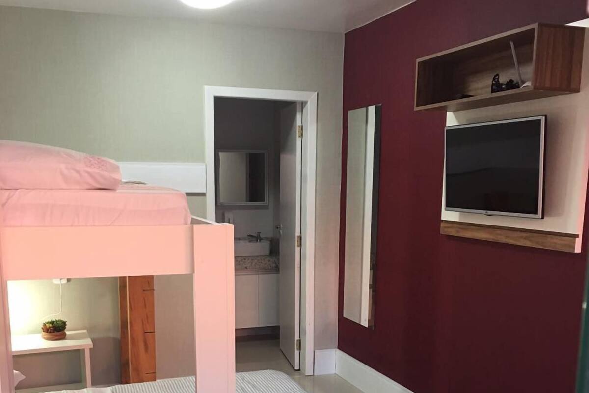 A bedroom with a bunk bed, a bedside table with a lamp, a TV mounted on the wall, and a bathroom visible through an open door.