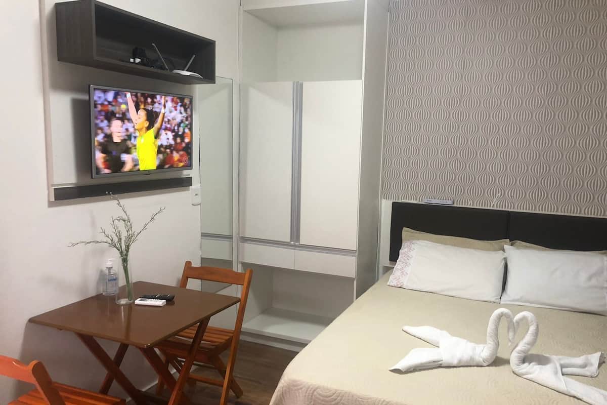 A hotel room with a bed, a TV mounted on the wall, a small table with a chair, and a decorative towel on the bed.